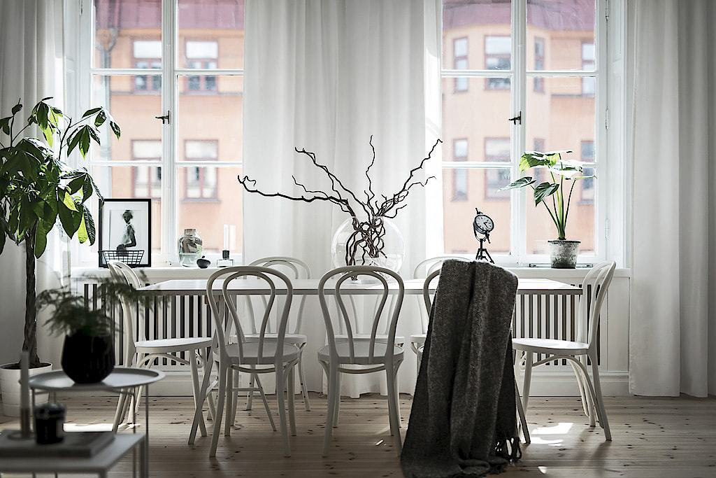 Hình ảnh bàn ghế ăn màu trắng đặt cạnh khung cửa sổ kính trong suốt, rèm cửa màu trắng, cây xanh trang trí