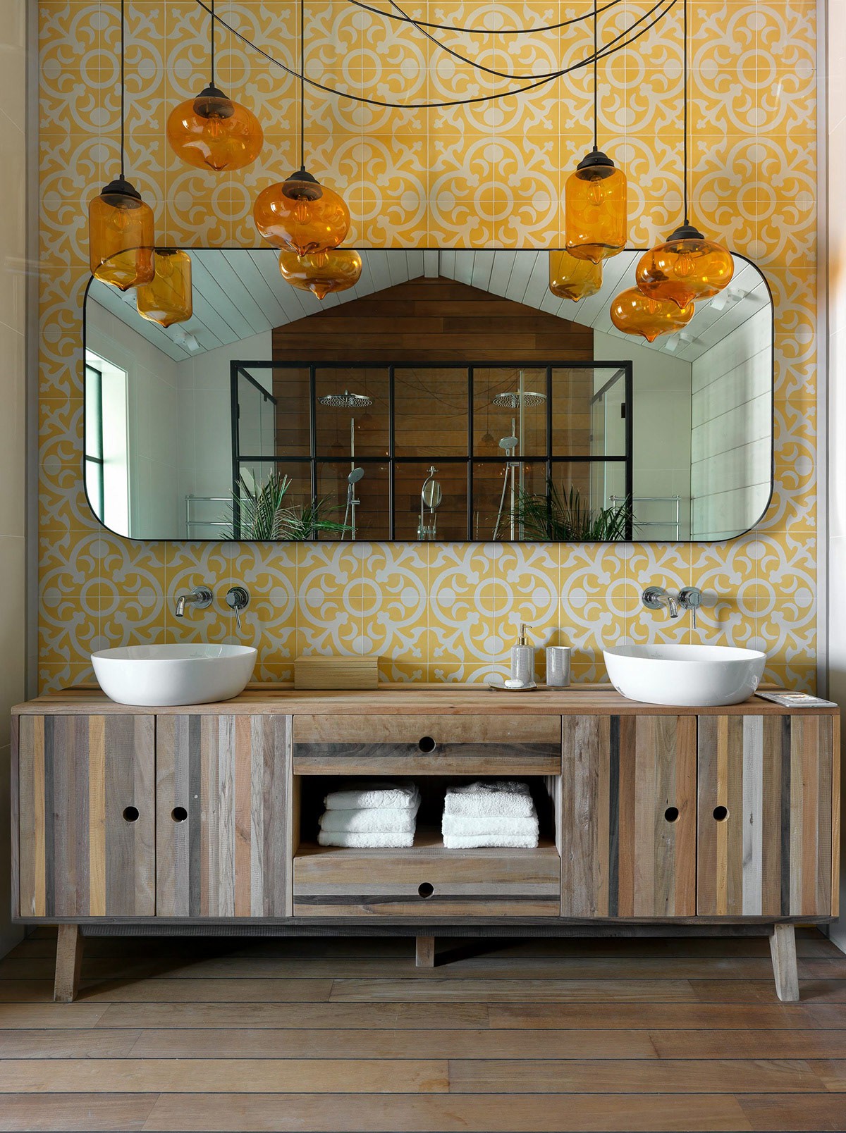 Hình ảnh phòng tắm nghệ thuật với giấy dán tường màu vàng chanh, bồn rửa bằng sứ trắng, đèn thả trang trí, tủ lưu trữ bằng gỗ cùng tông với sàn nhà