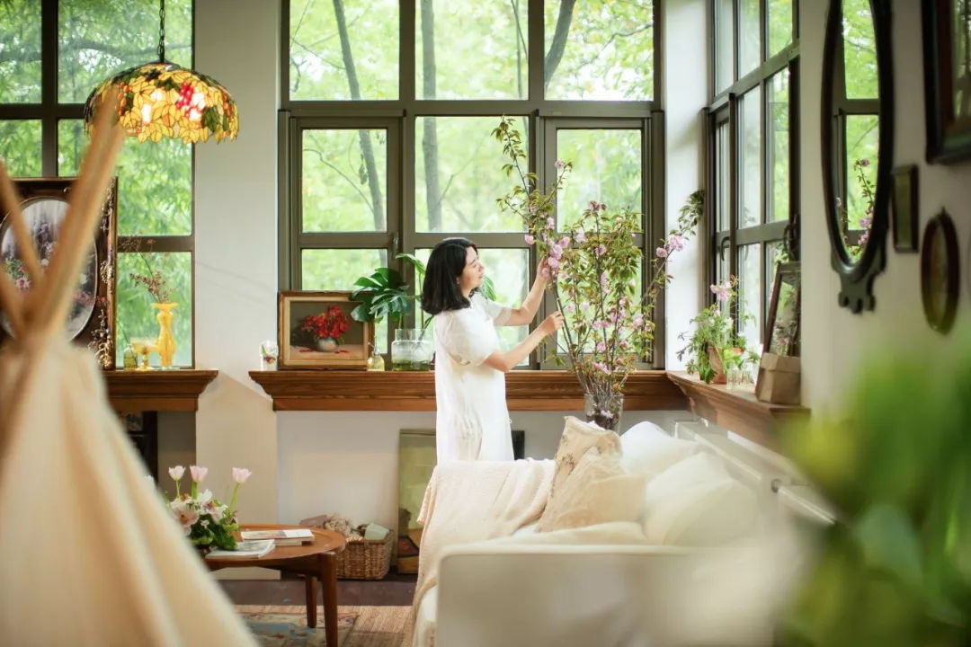 Hình ảnh một người phụ nữ đang cắm bình hoa trong phòng khách, nhiều khung cửa sổ kính, đèn trang trí màu sắc