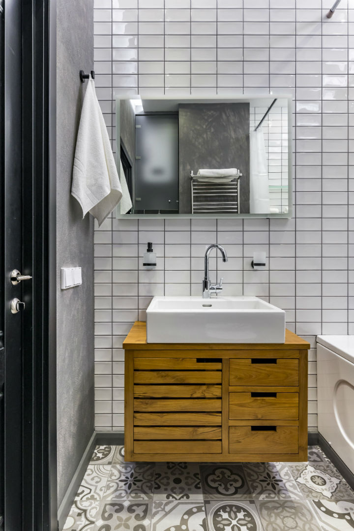 Hình ảnh phòng tắm với sàn lát gạch bông, tường ốp gạch trắng, gương soi, bồn rửa sứ trắng, tủ đồ bằng gỗ