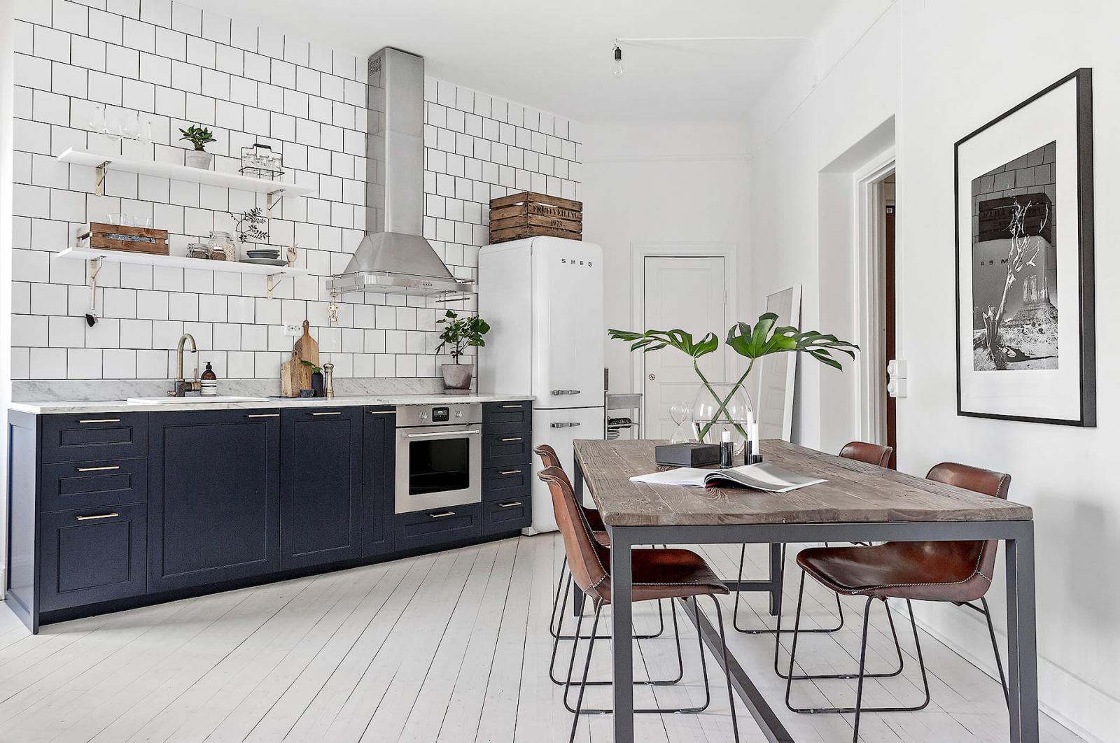 Hình ảnh toàn cảnh phòng bếp với tủ màu xanh than, tường ốp gạch trắng, máy hút mùi, tủ lạnh, bàn ăn bằng gỗ