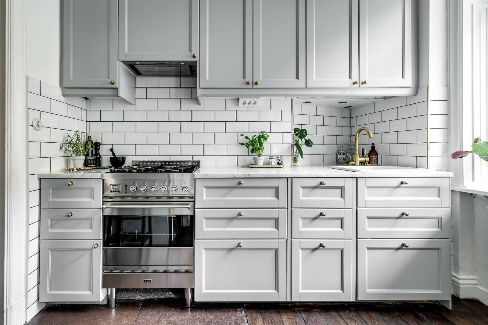 Hình ảnh cận cảnh hệ tủ bếp màu trắng xám gồm nhiều ngăn kéo, tay nắm kim loại, tường bếp ốp gạch trắng, cây xanh đặt trên bệ bếp