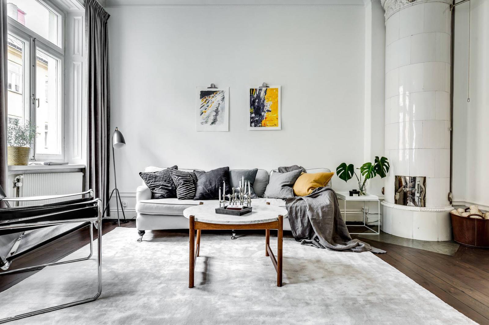 Hình ảnh toàn cảnh phòng khách với sofa trắng, tranh treo tường, cây xanh trang trí, gối tựa màu vàng chanh, cạnh đó là lò sưởi