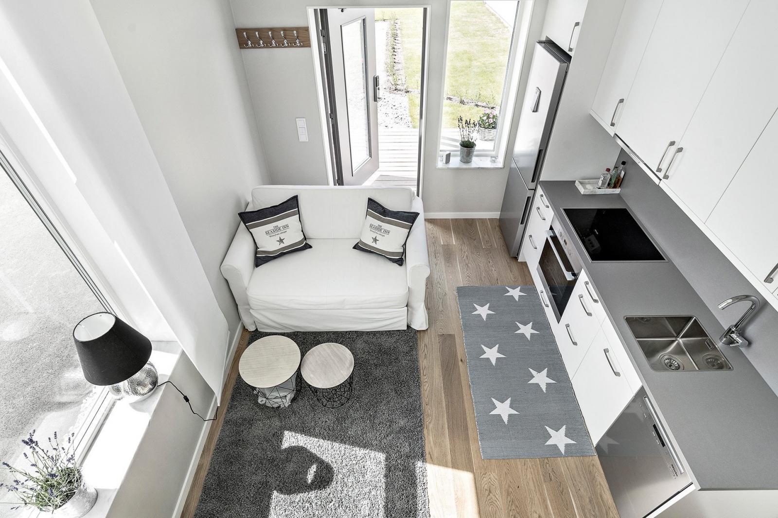 Hình ảnh toàn cảnh không gian sinh hoạt chung trong nhà 19m2 nhìn từ phòng ngủ xuống với sofa trắng, bếp nấu, thảm trài màu xám