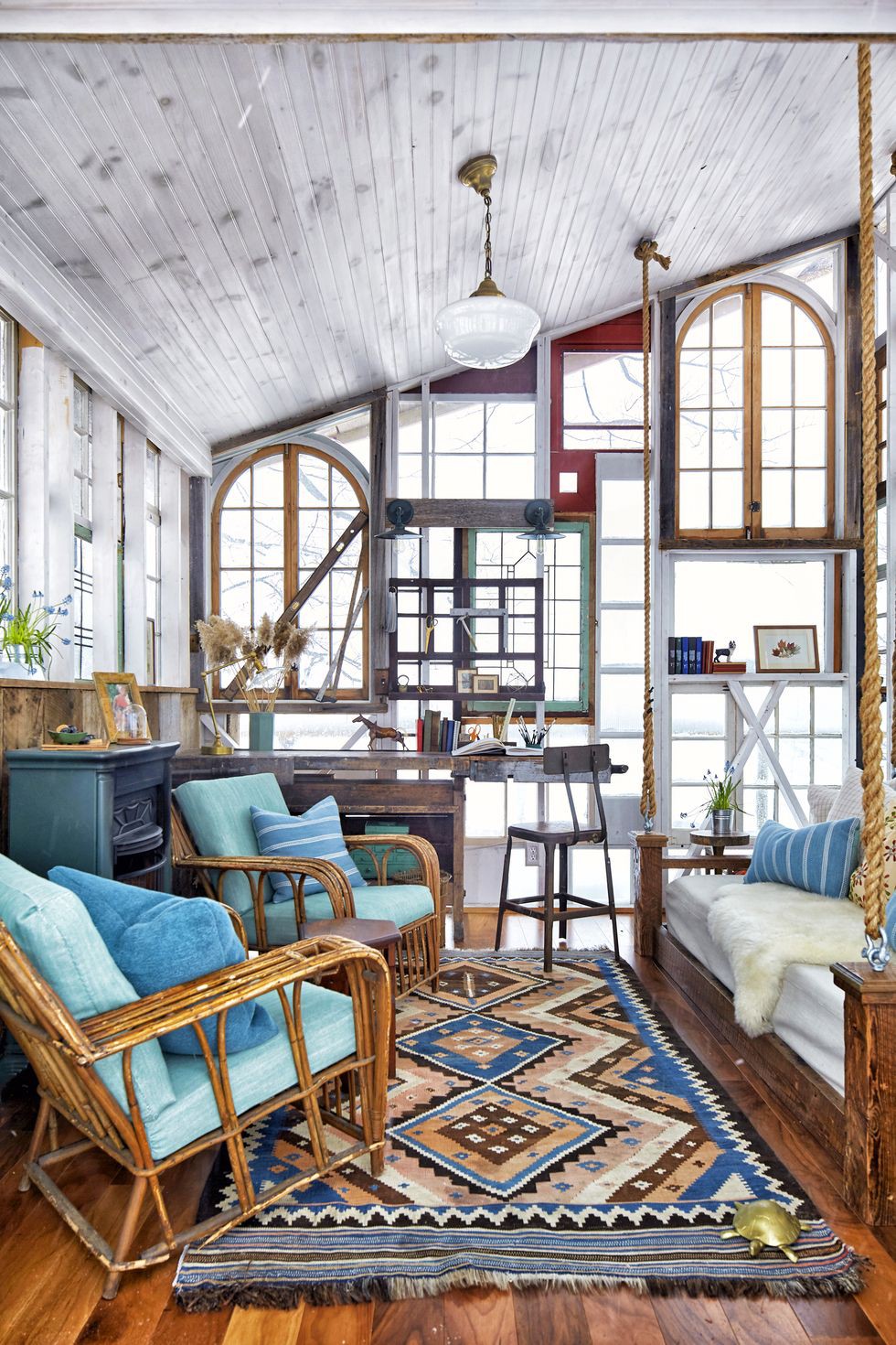 Hình ảnh bên trong ngôi nhà nhỏ làm từ khung cửa sổ cũ với ghế ngồi bằng mây lót nệm xanh dương, giường gỗ ngay cạnh, tủ kệ lưu trữ, bàn ghế, thảm trải sàn màu sắc