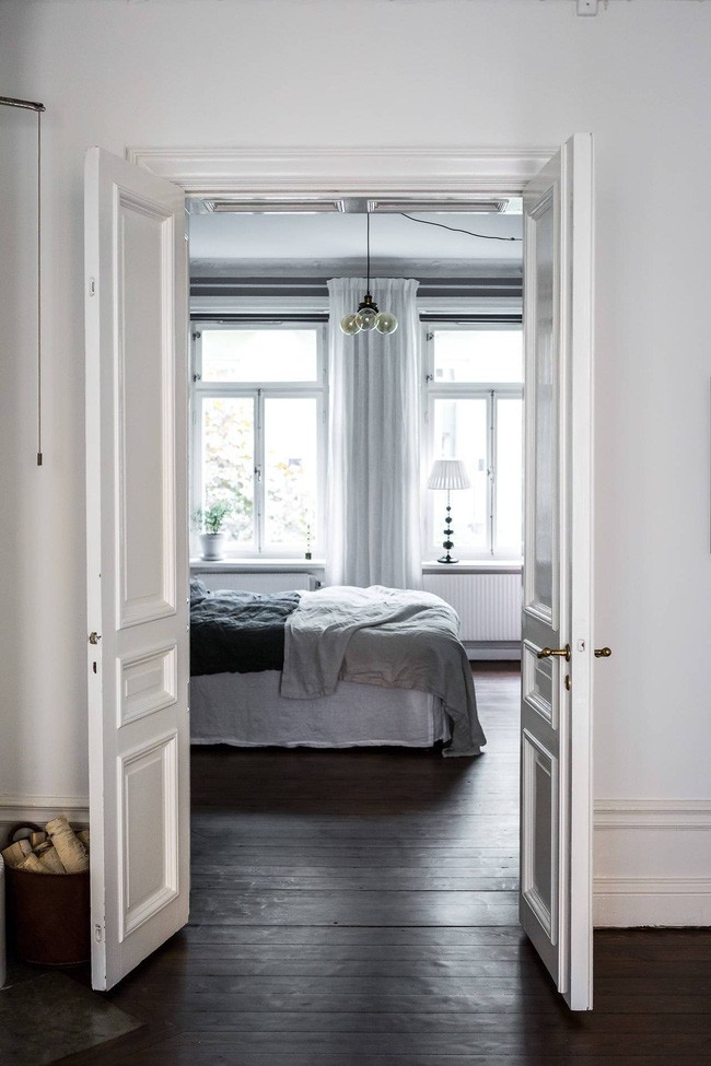 Hình ảnh phòng ngủ với ga gối màu trắng, cửa sổ kính nhìn từ phòng khách