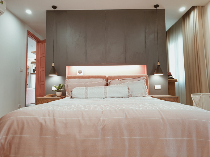 Hình ảnh cận cảnh phòng ngủ trong căn hộ 153m2 với ga gối màu hồng, tường đầu giường sơn xám đen, bộ đôi đèn thả 