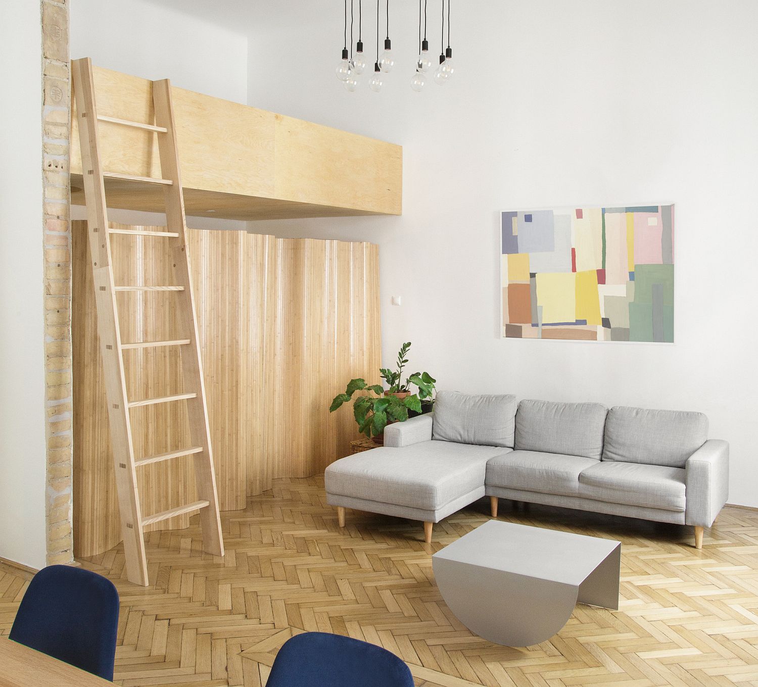 Phòng khách căn hộ có gác lửng được bài trí đơn giản với bộ ghế sofa màu xám sáng, bàn trà kim loại cùng tông; cây xanh, tranh tường tạo điểm nhấn.