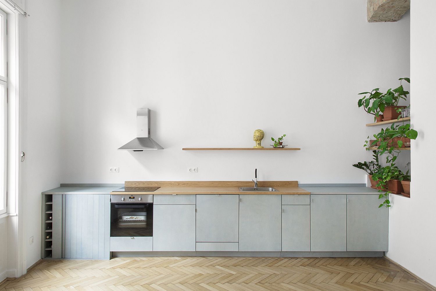 Hình ảnh phòng bếp màu trắng tối giản với tủ bếp dưới màu xám, ống hút mùi nhỏ, kệ cây xanh trang trí ở góc