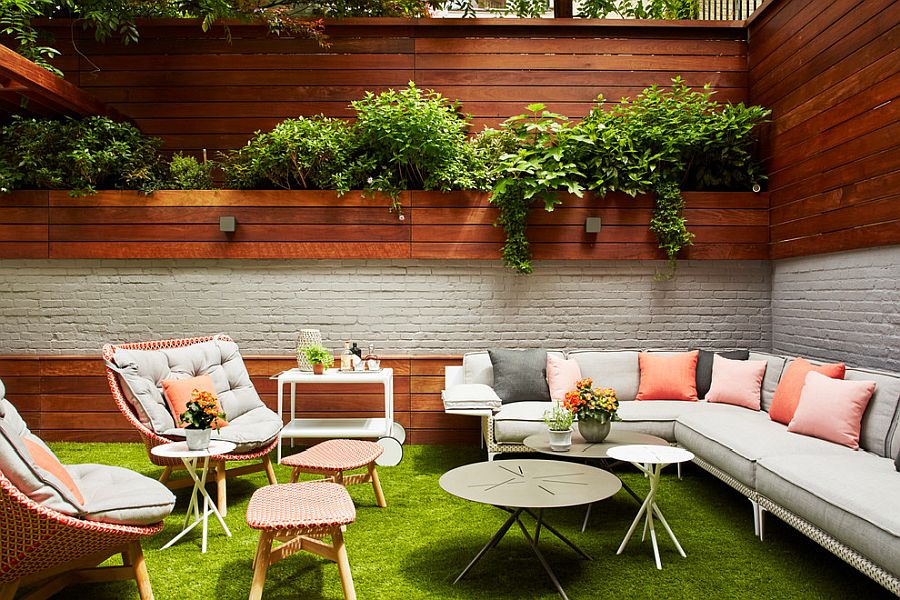 Hình ảnh không gian ngoài trời với bàn ghế thư giãn, gối tựa màu hồng,bàn trà đặt trên thảm cỏ, tường gỗ, cây xanh