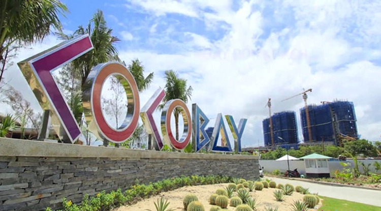 Hình ảnh lối vào dự án condotel Cocobay Đà Nẵng với biển tên màu tím, hàng dừa xanh tốt, cây bụi thấp, tòa nhà xây dở
