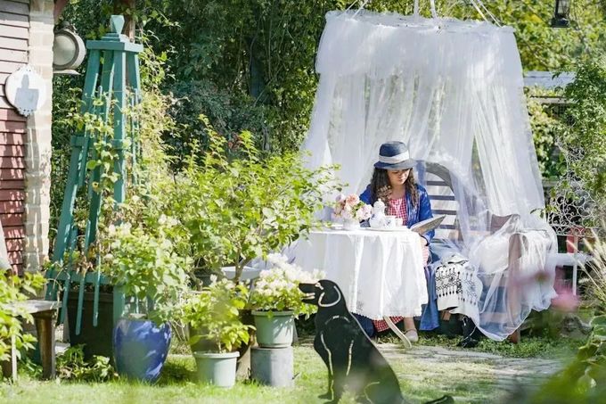 Hình ảnh một người phụ nữ đang đọc sách, làm việc trong vườn nhà, xung quanh là cây xanh, hoa cỏ, chú chó