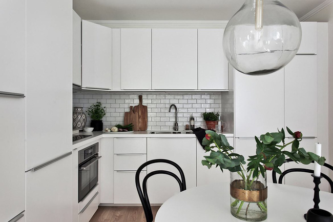 Hình ảnh cận cảnh phòng bếp với tủ đựng màu trắng, tường ốp gạch trắng, bàn ăn tròn, ghế đen, đèn thả