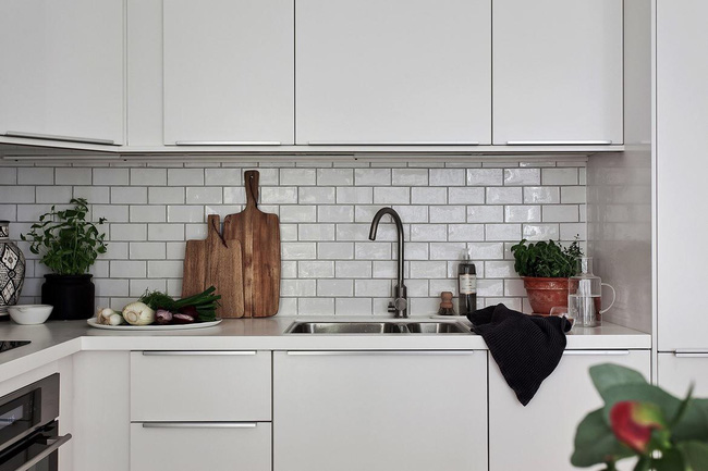 Hình ảnh cận cảnh một góc phòng bếp với tường ốp gạch, tủ bếp màu trắng, cây xanh, thớt gỗ