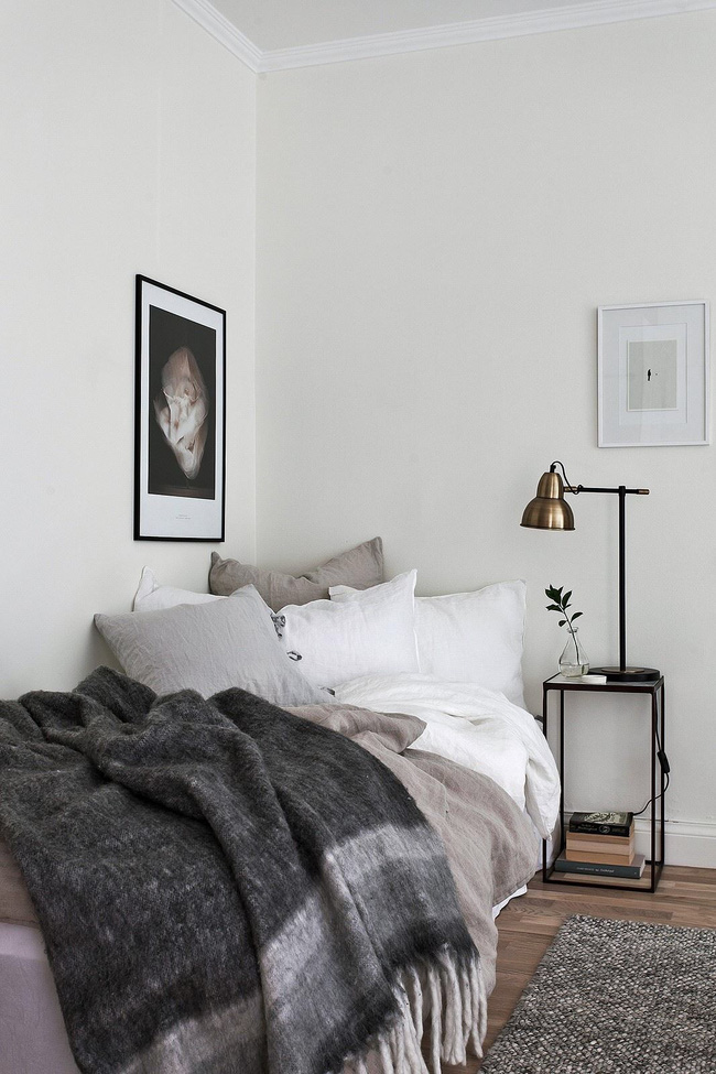 Hình ảnh cận cảnh giường ngủ với chăn gối màu xám - trắng, đèn đọc sách, tranh treo tường