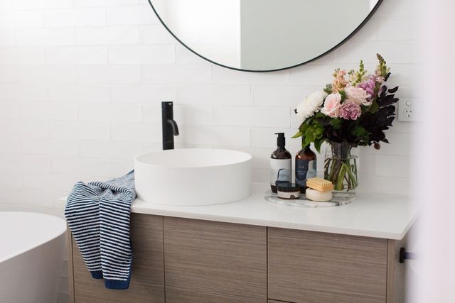 Hình ảnh một góc phòng tắm với gương tròn, bồn rửa bằng sứ trắng, vòi nước màu đen, lọ hoa trang trí, tinh dầu, tủ gỗ