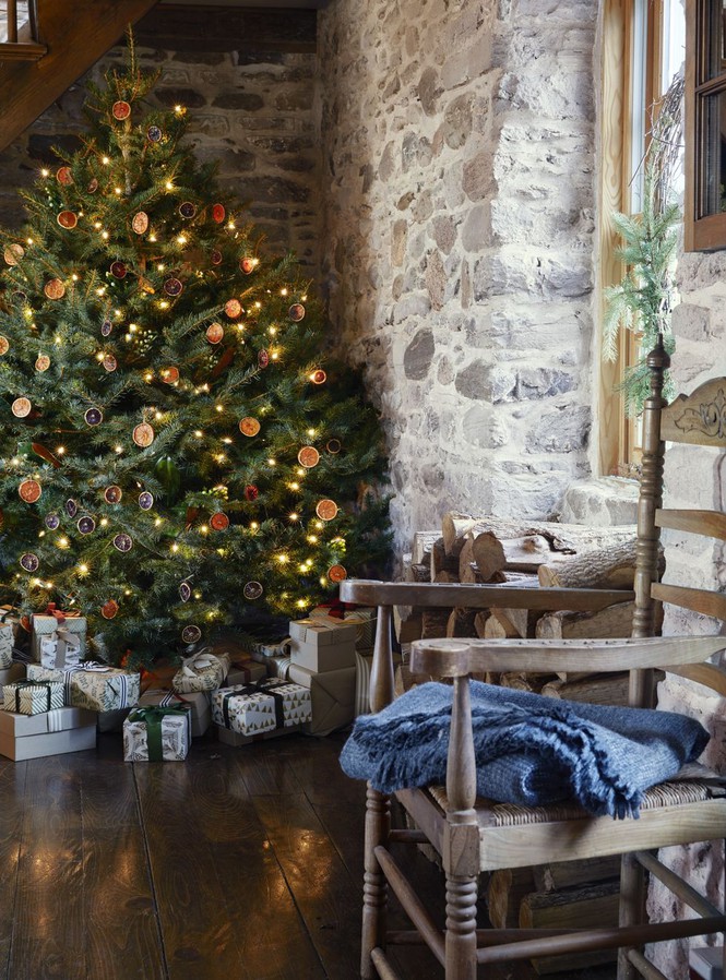 Hình ảnh cây thông Noel ở góc nhà được trang trí với phụ kiện màu đỏ, xanh, đèn LED, phía dưới gồm nhiều hộp quà, bên cạnh là ghế ngồi thư giãn