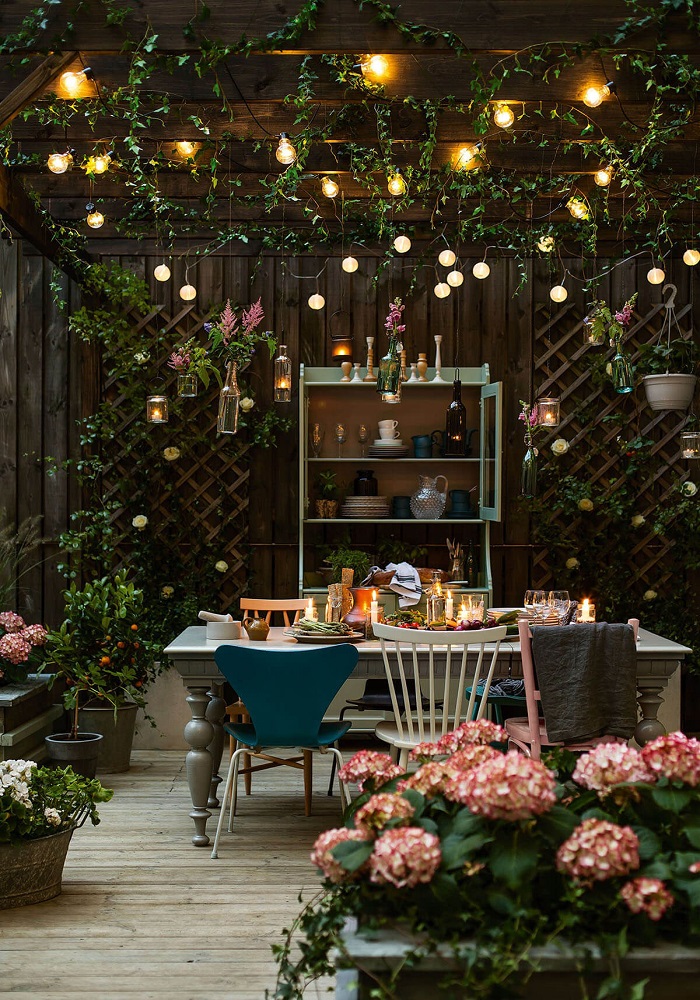Hình ảnh cận cảnh góc ăn uống, thư giãn nơi sân vườn được trang trí với tường gỗ, đèn LED, bàn ghế màu sắc, hoa hồng