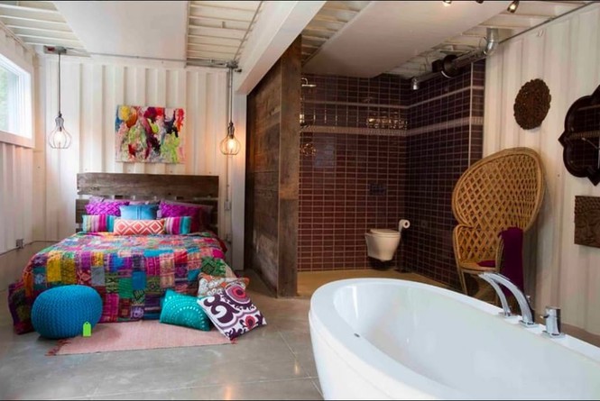 Hình ảnh một phòng ngủ bên trong nhà container với giường màu sắc, tranh trang trí, đèn thả sợi đốt đối xứng hai bên, cạnh đó là bồn tắm lớn