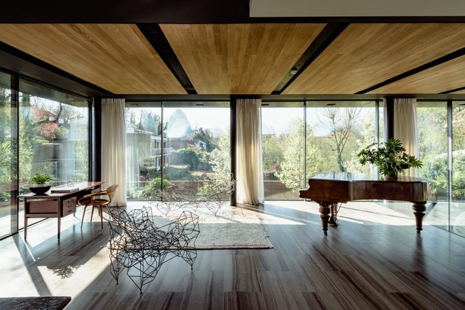 Hình ảnh một căn phòng rộng rãi có tường kính, trần ốp gỗ, đặt piano, bàn làm việc, cây xanh