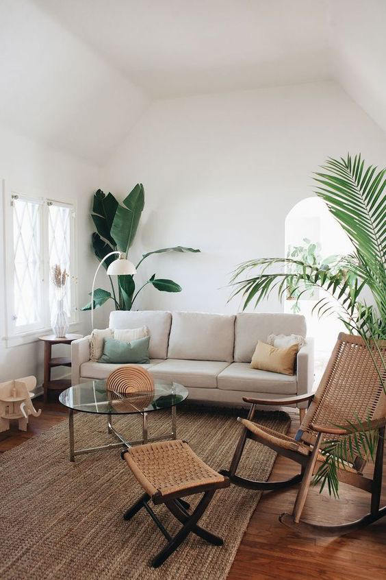 Hình ảnh một phòng khách có trần cao, tường màu trắng, sử dụng nội thất làm từ chất liệu tự nhiên như kệ gỗ, ghế ngồi mây tre đan, thảm cói dệt, cây xanh lớn trang trí ở góc phòng