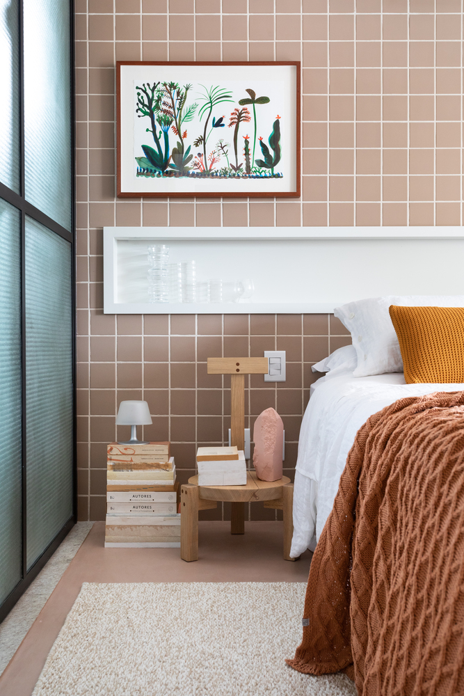 Hình ảnh cận cảnh một góc phòng ngủ với giường nệm cao êm ái, gạch ốp tường màu hồng đất, tranh trang trí, bàn gỗ đầu giường, vách kính mờ