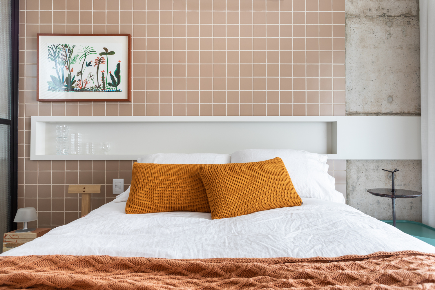 Hình ảnh một góc phòng ngủ với tường ốp gạch màu hồng đất, xen kẽ mảng tường bê tông, tranh treo tường, gối tựa màu vàng nổi bật