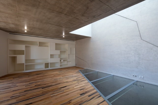 Hình ảnh một căn phòng trong biệt thự núi đa với tường bê tông xám, kệ gỗ màu trắng gắn tường, một phần sàn lát kính trong suốt