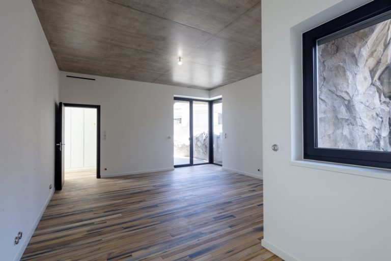 Hình ảnh một căn phòng trống với sàn lát gỗ, tường sơn trắng, tường bê tông xám, cửa kính trong suốt
