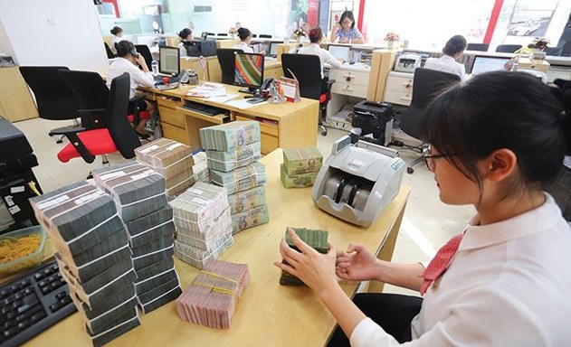 Hình ảnh cận cảnh một nhân viên ngân hàng đang kiểm đếm các cọc tiền trước mặt, xung quanh nhiều người đang làm việc