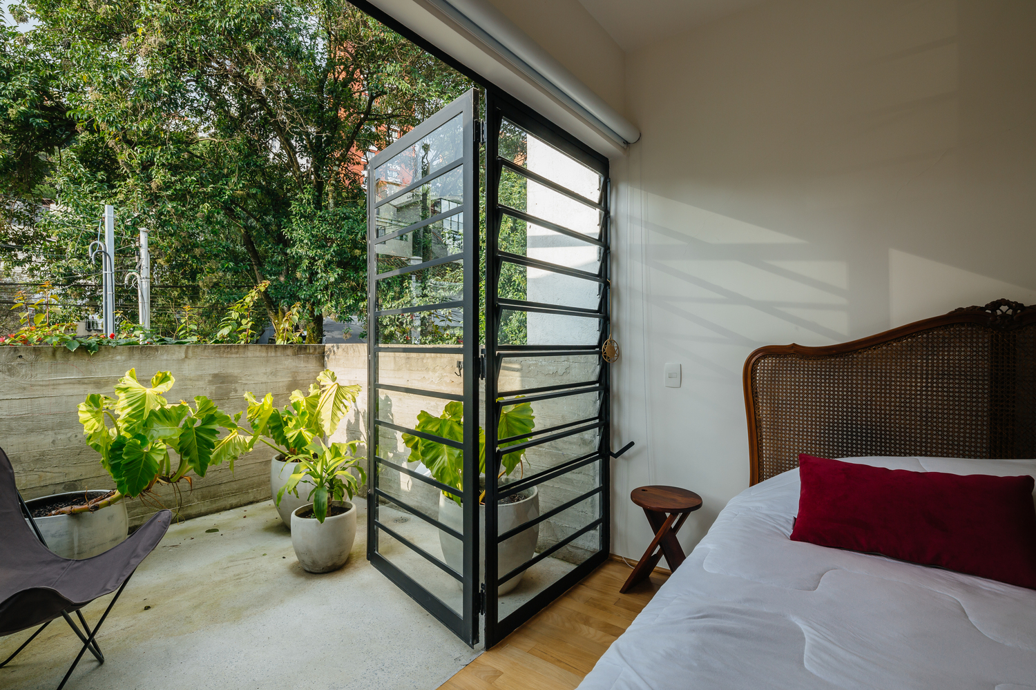 Hình ảnh khung cửa kính phòng ngủ mở ra ban công trồng cây xanh, đặt ghế ngồi thư giãn, tường bê tông