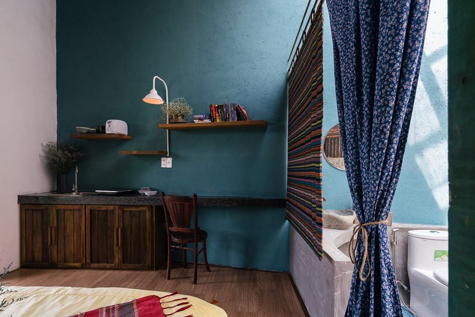 Hình ảnh cận cảnh góc nấu nướng, đọc sách trong phòng nghỉ dưỡng với bức tường sơn màu xanh dương, kệ gỗ gắn tường, rèm thổ cẩm, hoa nhí