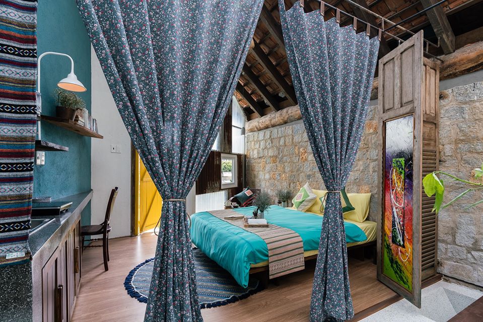 Hình ảnh toàn cảnh phòng ngủ trong nhà nghỉ dưỡng nhìn từ khu vệ sinh với rèm cửa hoa nhí, tường sơn xanh, kệ gỗ gắn tường, giường nệm êm ái