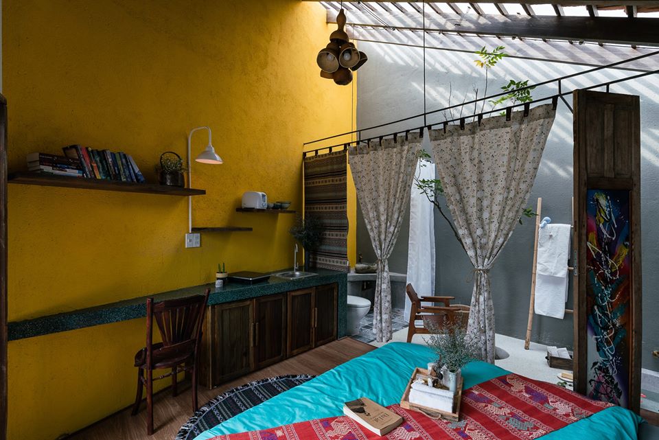 Hình ảnh một góc phòng ngủ với bức tường sơn màu vàng chanh ấn tượng, góc đọc sách, nấu nướng nhỏ gọn, giường nệm màu xanh dương chủ đạo, rèm cửa Vintage lãng mạn