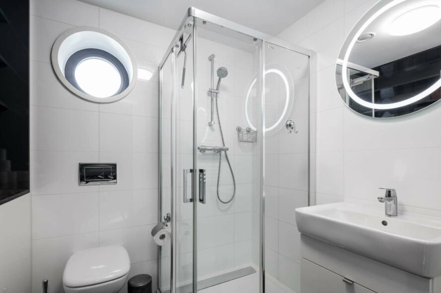 Hình ảnh phòng tắm tông màu trắng chủ đạo với góc tắm đứng phân tách bằng vách kính trong suốt, cửa sổ tròn phía trên lấy sáng