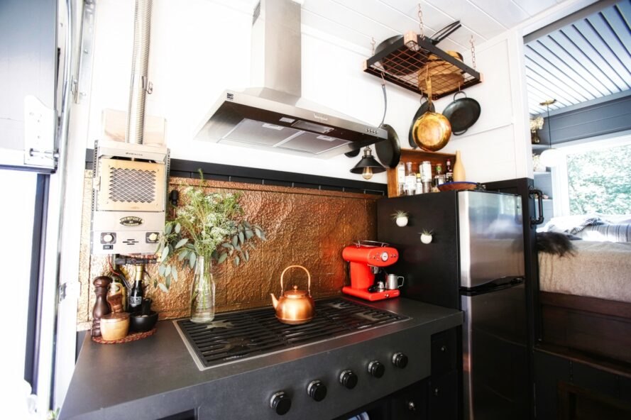 Hình ảnh phòng bếp nhỏ hiện đại với giá treo soong chảo gắn tường, tường chắn ốp đồng, cây xanh trang trí