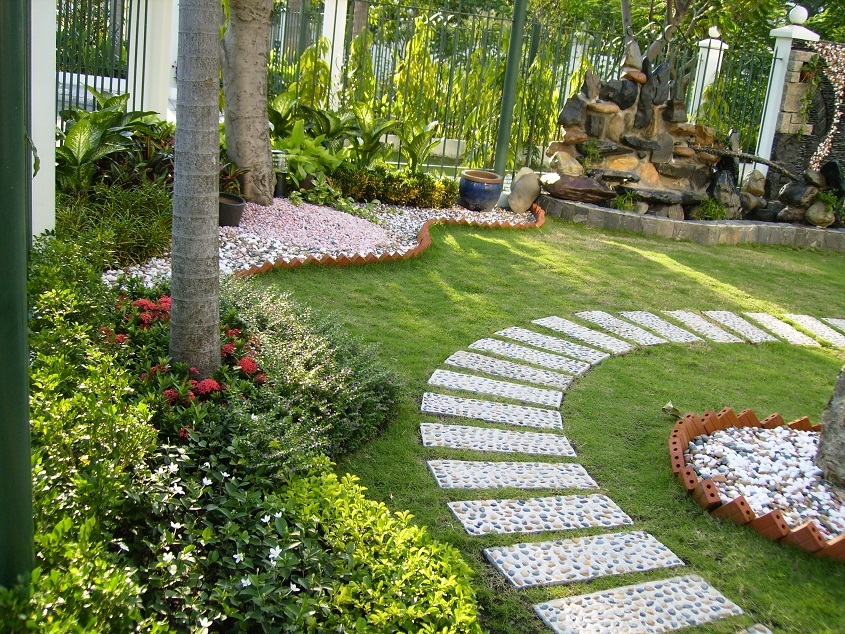 Hình ảnh lối đi trong khu vườn được làm bằng sỏi kết hợp xi măng, hai bên trồng cỏ, nhiều cây lớn bao quanh.