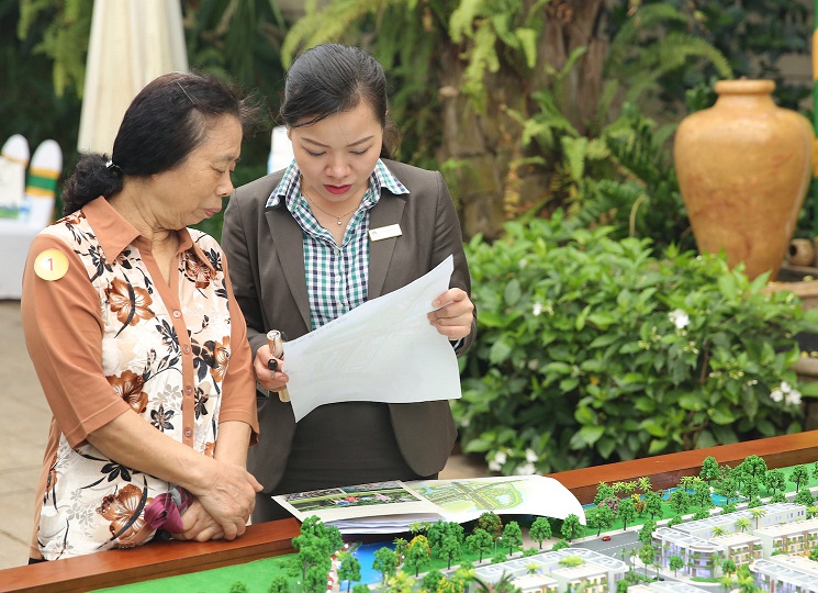 Hình ảnh nữ nhân viên môi giới đang giới thiệu dự án cho khách hàng nữ luống tuổi, cạnh đó là sa bàn dự án
