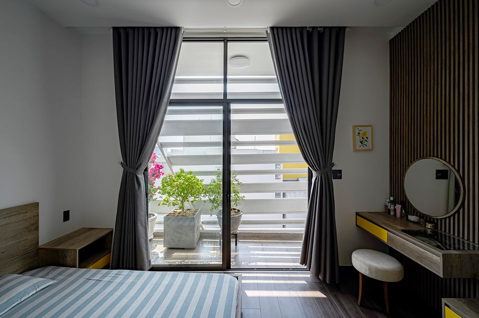 Hình ảnh một góc phòng ngủ với giường nệm kẻ sọc, bàn trang điểm, cửa kính lớn mở ra ban công trồng hoa, cây xanh.