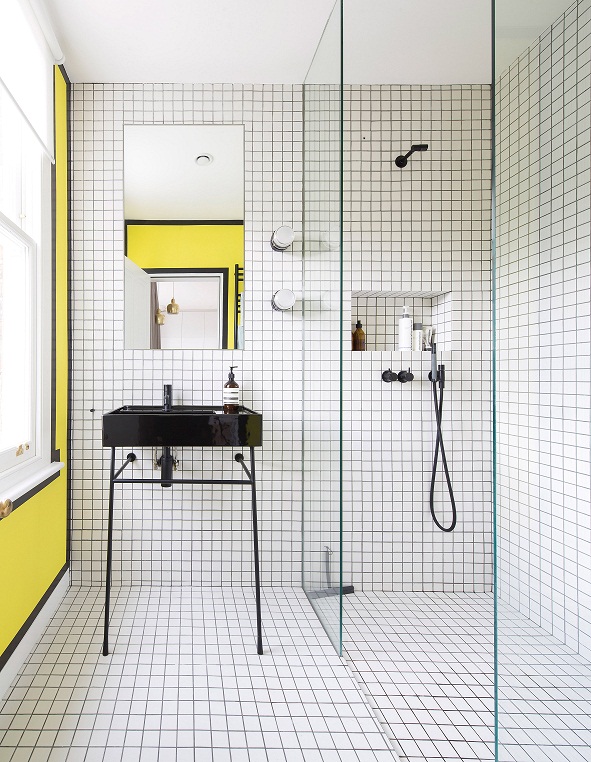 Hình ảnh một phòng tắm nhỏ với tường và sàn ốp gạch men màu trắng cỡ nhỏ, điểm nhấn màu vàng chanh từ cửa sổ, buồng tắm kính