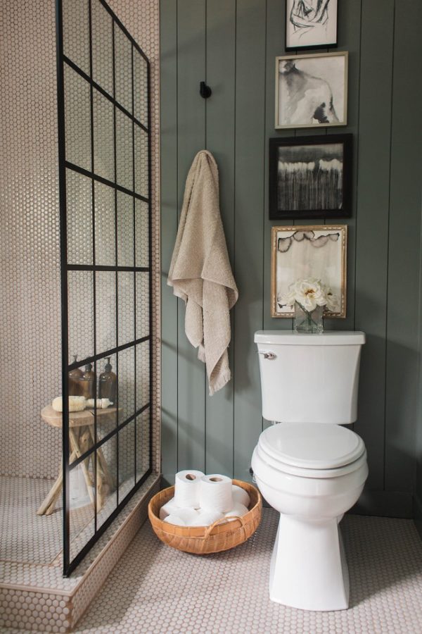 Hình ảnh cận cảnh một góc phòng tắm với mảng tường phía sau bồn cầu ốp gỗ sơn xanh, tường buồng tắm ốp gạch họa tiết tổ ong, giữa là vách ngăn kính khung đen