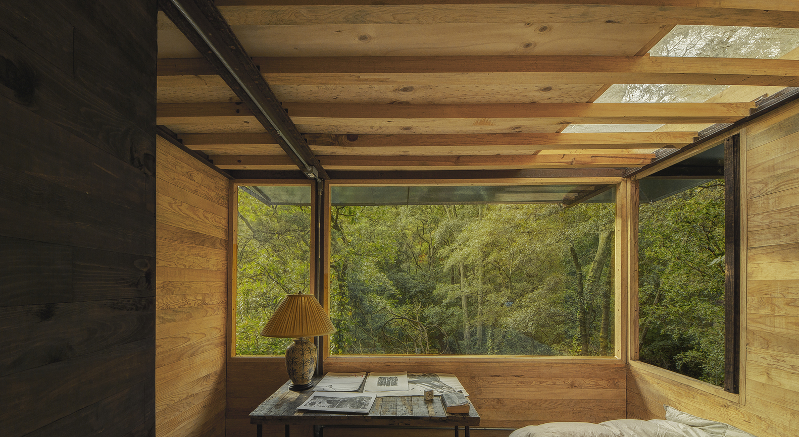 Hình ảnh một góc phòng ngủ với bàn gỗ đặt đèn chụp, cửa kính lớn nhìn ra rừng cây xanh