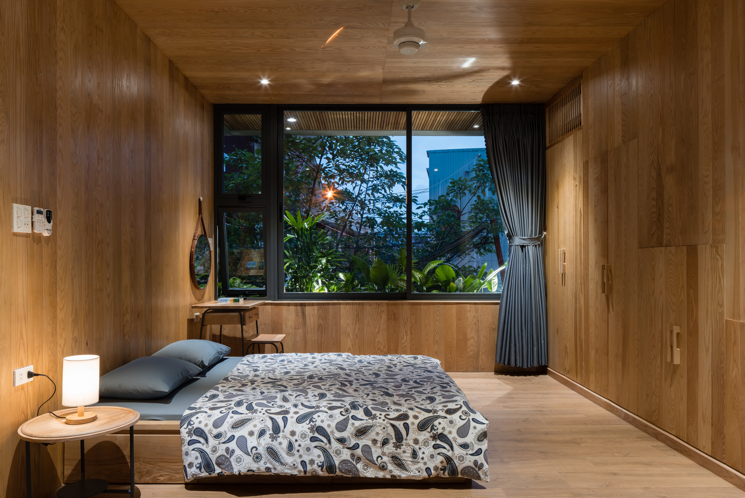 Hình ảnh toàn cảnh phòng ngủ lớn với chất liệu gỗ chủ đạo, bàn tròn đầu giường, đèn âm trần, cửa kính lớn mở ra sân vườn