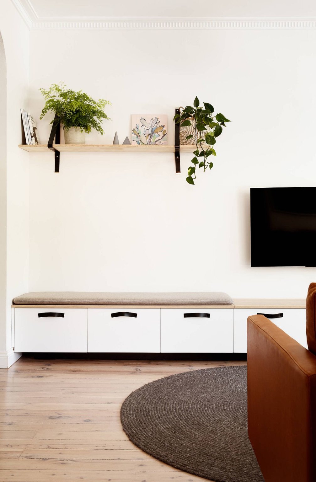 Hình ảnh một góc phòng khách nhỏ với tường màu trắng làm nền cho kệ gỗ, cây xanh, sofa nâu da bò nổi bật hơn