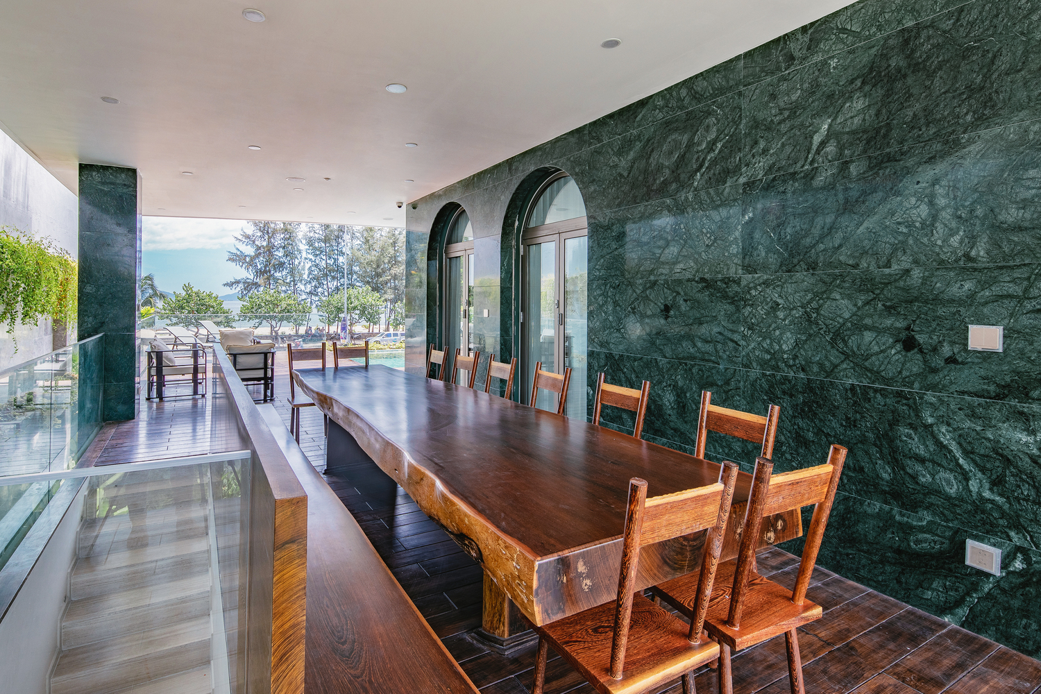 Hình ảnh cận cảnh bàn ăn dài làm bằng gỗ nguyên khối trong biệt thự, cạnh đó là tường ốp đá màu xanh ngọc sang trọng