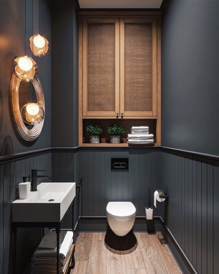 Hình ảnh phòng tắm hiện đại với tường màu xám đen, thiết bị màu trắng, tủ gỗ lưu trữ, gương tròn, đèn vàng ấm áp.