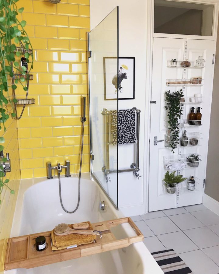Hình ảnh cận cảnh một góc phòng tắm với tường ốp gạch màu vàng chanh ngay trên bồn tắm nắm, phía ngoài vách kính là giá treo tường, trang trí chậu cây xanh.