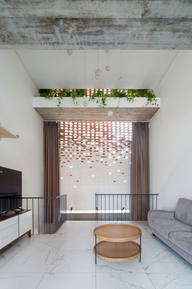 Hình ảnh phòng khách có thiết kế đơn giản với sofa màu da bò, bàn trà tròn bằng gỗ, đối diện là tủ kệ tivi, rèm cửa mở ra lớp tường gạch nung
