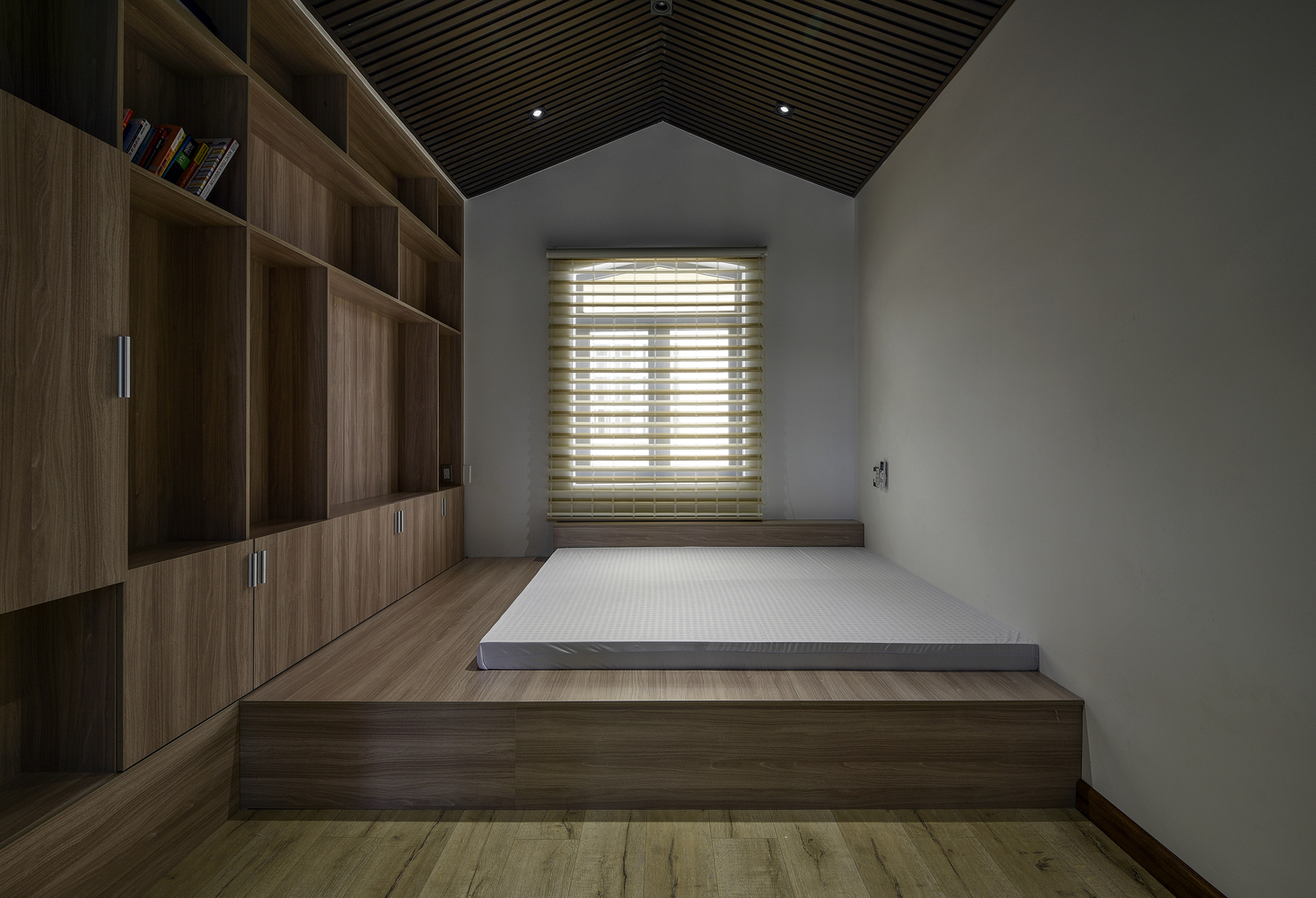 Hình ảnh toàn cảnh phòng ngủ tối giản với nệm trắng đặt trên bục gỗ cao hơn mặt sàn, cạnh dó là hệ tủ lưu trữ cao kịch trần kiểu mở 