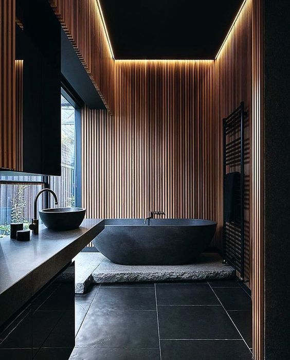 Hình ảnh toàn cảnh phòng tắm phong cách mở tông màu đen chủ đạo với tường ốp gỗ thanh dọc màu sáng, chậu rửa và bồn tắm đá tạo điểm nhấn